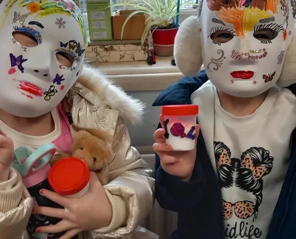 Two children wearing decorated white masks and holding small containers, with one child on the left also holding a water bottle.